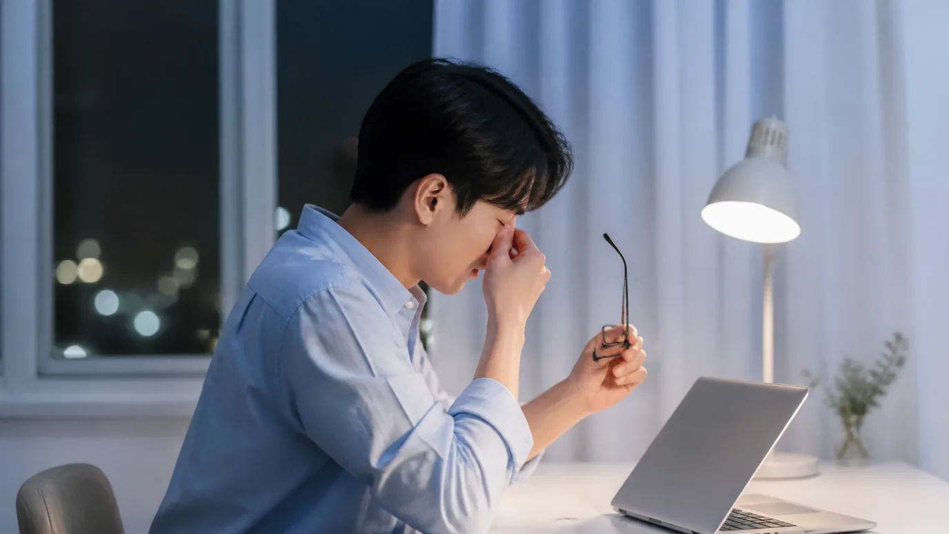 밤 책상 앞에서 안경을 벗고 눈가를 짚은 남성 — 눈의 피로