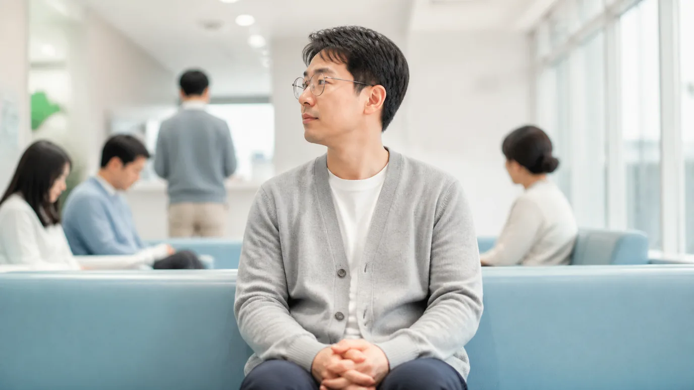 병원 대기 공간에서 30대 한국인 성인이 안경을 쓴 채 두 손을 편안히 모으고 기다리는 장면