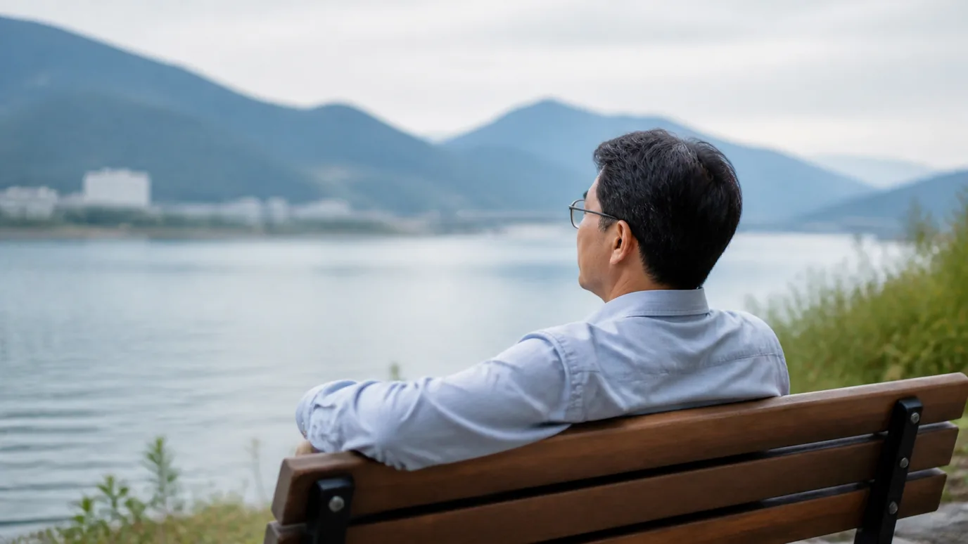 강변 산책로 벤치에서 50대 한국인 성인이 안경을 쓴 채 먼 풍경을 바라보는 장면