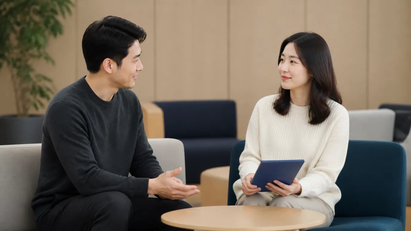 조용한 사무실 휴게 공간에서 30대 한국인 성인이 닫힌 단색 종이 복지 책자를 무릎에 놓고 동료와 이야기하는 장면
