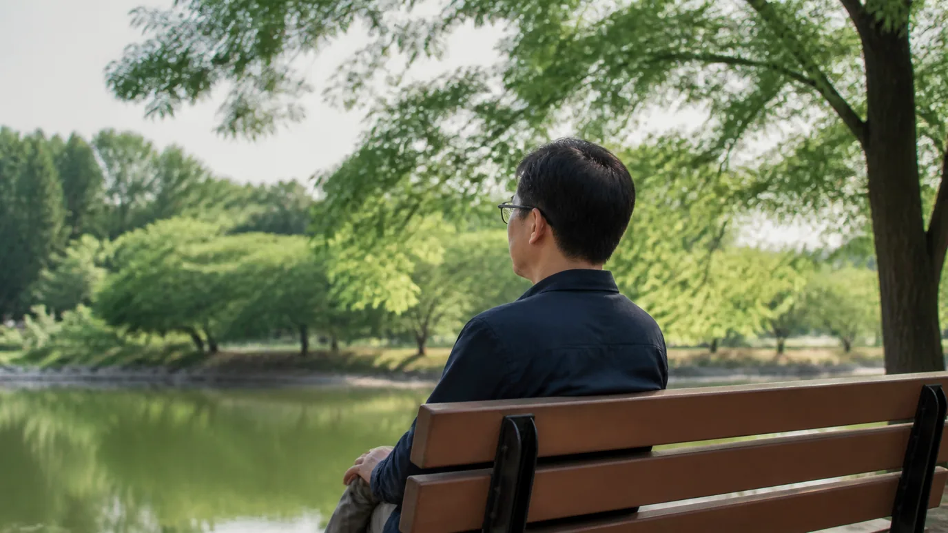 공원 벤치에서 40대 한국인 성인이 안경을 쓴 채 먼 나무를 편안히 바라보는 장면